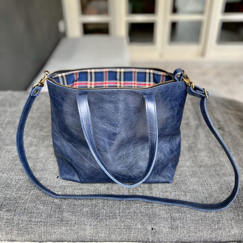 May include: A blue leather tote bag with a plaid fabric lining and a long adjustable strap. The bag has two top handles.