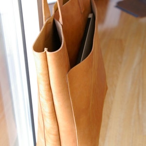 May include: A large, tan leather tote bag with a zippered pocket on the front. The bag is made of a soft, supple leather and has a spacious interior.