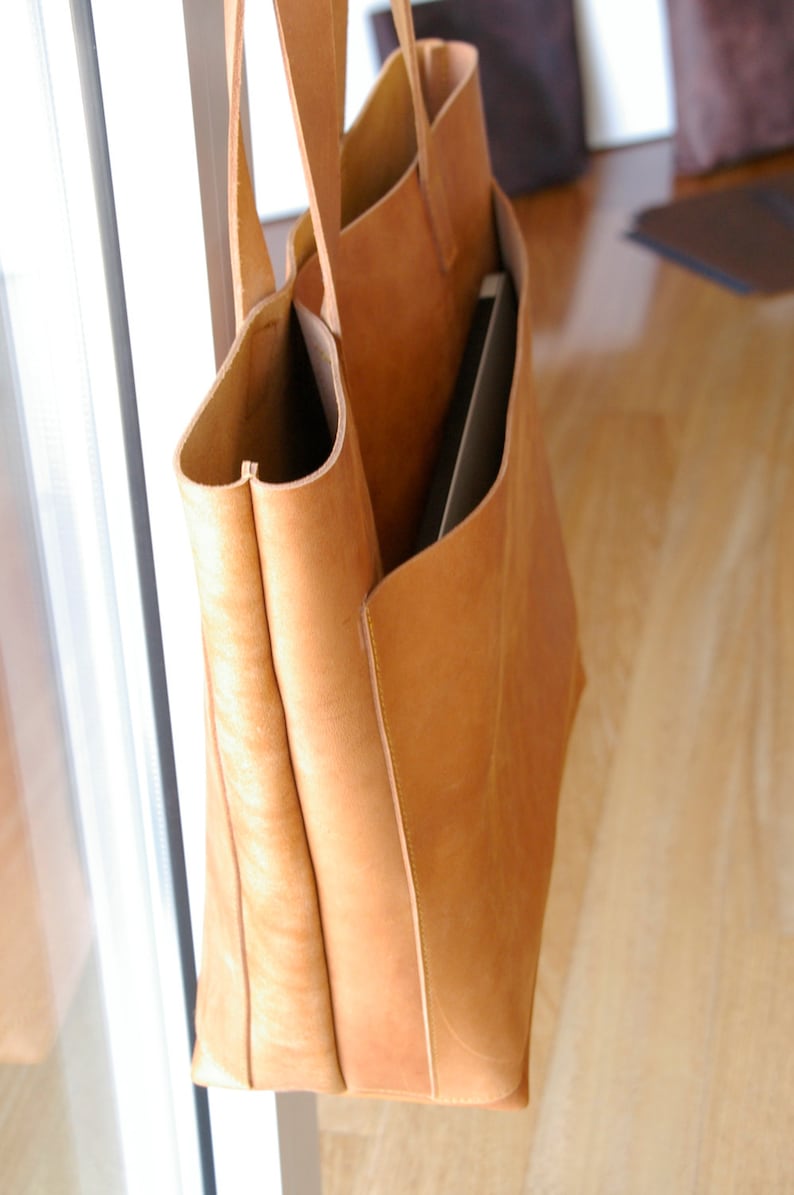 May include: A large, tan leather tote bag with a zippered pocket on the front. The bag is made of a soft, supple leather and has a spacious interior.