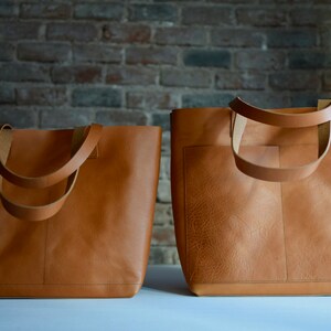 May include: Two brown leather tote bags, one slightly larger than the other, are displayed on a white surface. Both bags have a single top handle and a large open top.