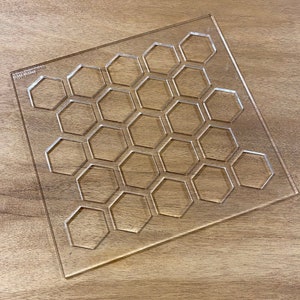 May include: Transparent acrylic template featuring a hexagonal grid pattern. The rectangular template has rounded corners and is placed on a wooden surface. The text "Artisan Model Makers" and a phone number are printed on the template.