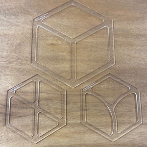 May include: Three clear plastic stencils for crafting, each shaped like a hexagon with different internal shapes. The stencils are on a wooden surface. The text "Artisan Model Makers 01143 803948" is visible on two of the stencils.