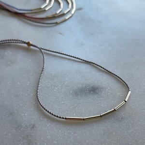 May include: A delicate gray cord necklace with gold-colored cylindrical beads. Several similar necklaces are visible in the background, with cords in shades of pink, blue, and gray. The jewelry rests on a white marble surface.