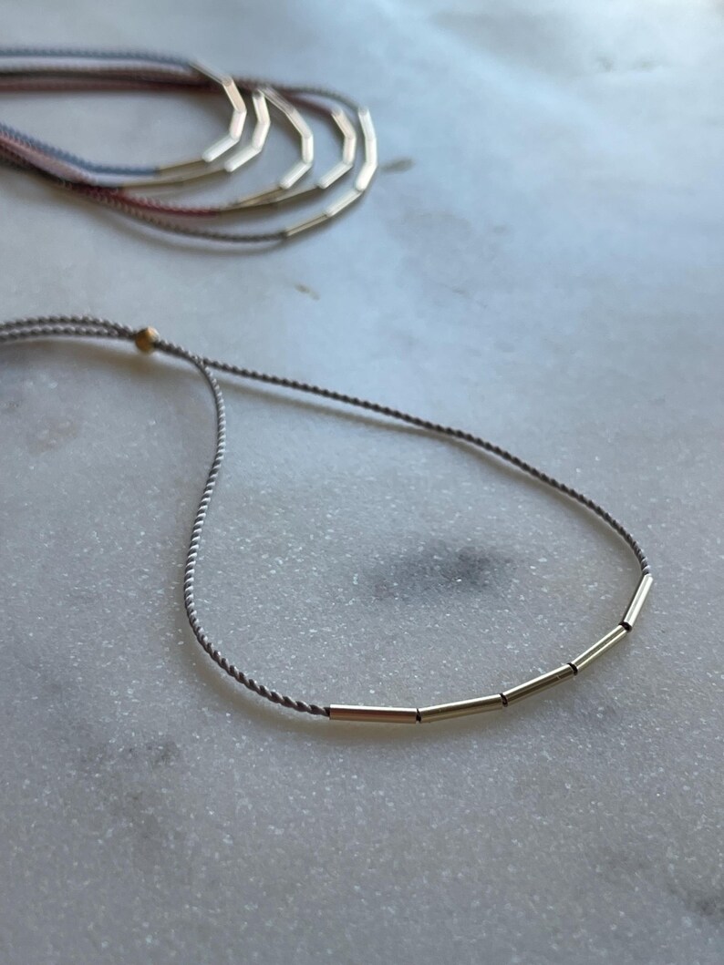 May include: A delicate gray cord necklace with gold-colored cylindrical beads. Several similar necklaces are visible in the background, with cords in shades of pink, blue, and gray. The jewelry rests on a white marble surface.