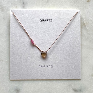 May include: A delicate necklace featuring a small, faceted, smoky quartz pendant and pink gemstone beads. The necklace is strung on a thin, brown cord and displayed on a white card with the words "QUARTZ" and "healing".