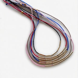 May include: A collection of colorful string necklaces with gold-colored cylindrical beads. The strings are in various colors, including red, black, blue, pink, and brown. The necklaces are displayed on a white background.