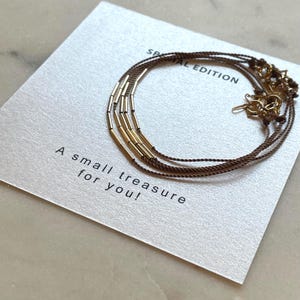 May include: A gold-toned bracelet with cylindrical beads and brown cord is displayed on a card. The card reads "Special Edition" and "A small treasure for you!" in black text. The bracelet is a delicate piece of jewelry.
