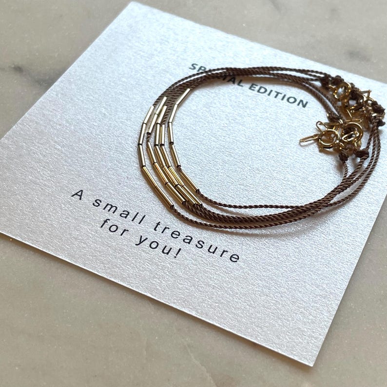May include: A gold-toned bracelet with cylindrical beads and brown cord is displayed on a card. The card reads "Special Edition" and "A small treasure for you!" in black text. The bracelet is a delicate piece of jewelry.