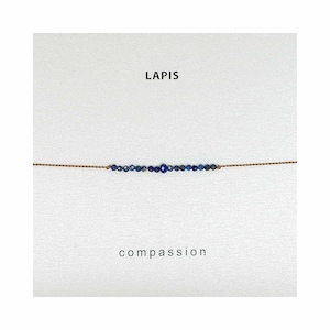 May include: A delicate bracelet with a single row of small, dark blue lapis lazuli beads strung on a thin brown cord. The bracelet is displayed on a white background with the words "LAPIS" and "compassion" printed below it.