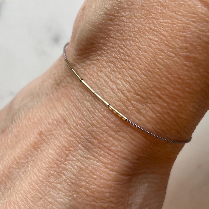 May include: A delicate gold bracelet with a minimalist design. The bracelet features several small gold cylindrical beads strung on a thin, gray cord. The bracelet is shown on a wrist.