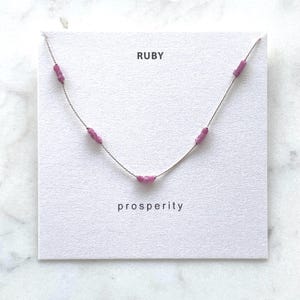 May include: A delicate silver-toned necklace featuring five small, cylindrical, magenta-colored ruby beads spaced along the chain. The necklace is displayed on a white card with the text RUBY printed at the top and prosperity printed at the bottom.