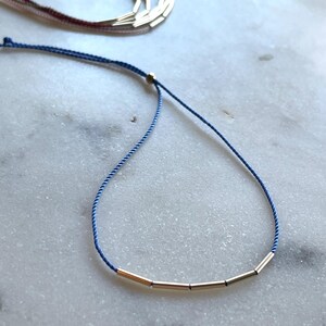 May include: A delicate gold-toned necklace with a blue cord. The necklace features several small, cylindrical gold beads strung along the cord. Additional necklaces are visible in the background, suggesting a jewelry collection.
