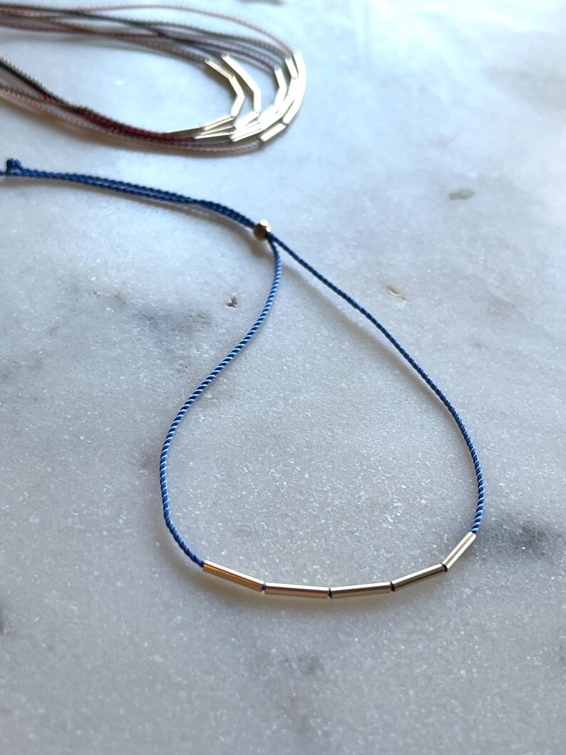 May include: A delicate gold-toned necklace with a blue cord. The necklace features several small, cylindrical gold beads strung along the cord. Additional necklaces are visible in the background, suggesting a jewelry collection.