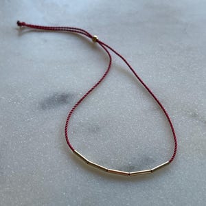 May include: A minimalist bracelet featuring four gold-colored cylindrical beads strung on a deep red cord. The adjustable cord allows for a customized fit. The bracelet is displayed on a white and gray marble surface.