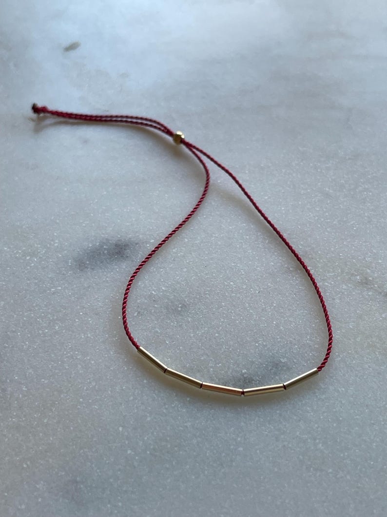 May include: A minimalist bracelet featuring four gold-colored cylindrical beads strung on a deep red cord. The adjustable cord allows for a customized fit. The bracelet is displayed on a white and gray marble surface.