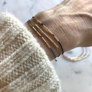 May include: Three delicate bracelets with gold-colored cylindrical beads and thin cords in black, blue, and gold. The bracelets are worn on a wrist, with a cream-colored sweater sleeve in the foreground. Additional bracelets are visible in the background.