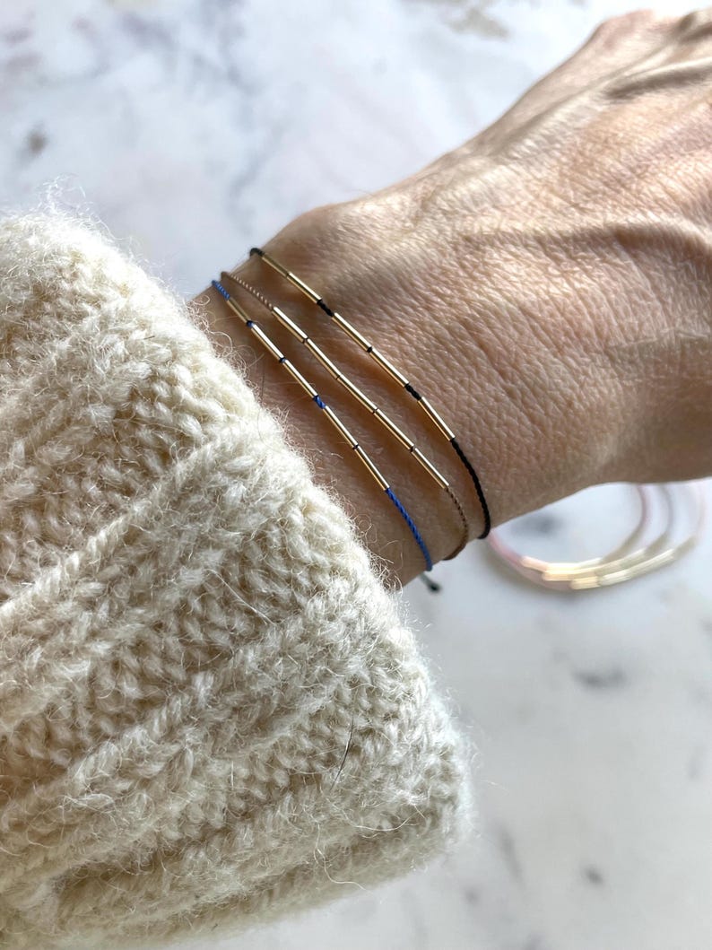 May include: Three delicate bracelets with gold-colored cylindrical beads and thin cords in black, blue, and gold. The bracelets are worn on a wrist, with a cream-colored sweater sleeve in the foreground. Additional bracelets are visible in the background.