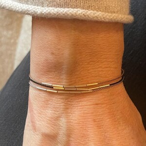 May include: A close-up of a wrist adorned with three delicate bracelets. The bracelets feature thin, dark cords with gold-colored cylindrical beads. The wrist is against a neutral-toned background, with a portion of a sweater visible.