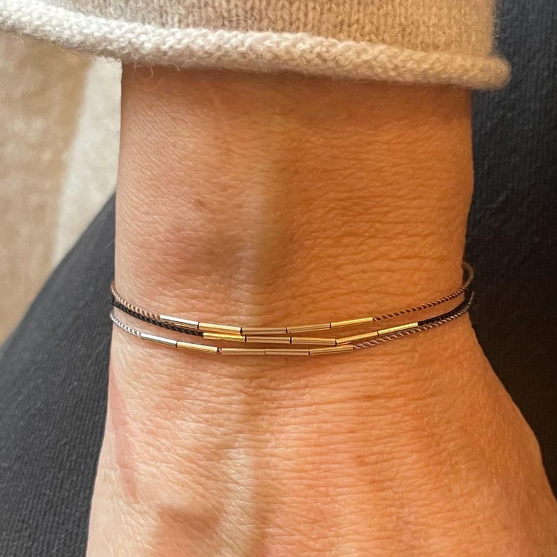 May include: A close-up of a wrist adorned with three delicate bracelets. The bracelets feature thin, dark cords with gold-colored cylindrical beads. The wrist is against a neutral-toned background, with a portion of a sweater visible.
