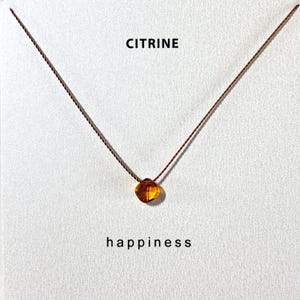 May include: A delicate necklace featuring a small, round, amber-colored citrine gemstone pendant. The pendant hangs from a thin, brown cord. The word "CITRINE" is printed above the pendant, and the word "happiness" is printed below.