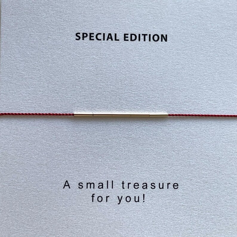 May include: A minimalist bracelet featuring a thin gold bar centered on a red cord. The text "SPECIAL EDITION" is printed above the bracelet, and the text "A small treasure for you!" is printed below.