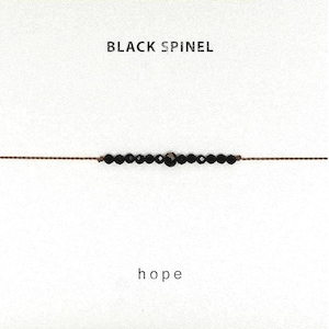 Peut inclure: Un bracelet délicat composé d'une rangée de petites perles de spinelle noir à facettes enfilées sur un fin cordon marron. Le texte "BLACK SPINEL" est imprimé au-dessus du bracelet et le mot "hope" est imprimé en dessous sur un fond blanc.