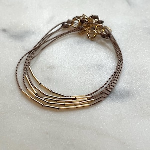May include: A set of three delicate bracelets. Each bracelet features a thin, brown cord adorned with gold-colored cylindrical beads. The bracelets are connected with gold-colored clasps, creating a layered look. The background is a light, marbled surface.