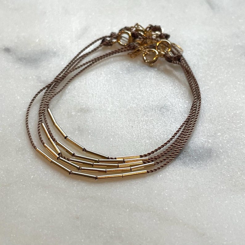 May include: A set of three delicate bracelets. Each bracelet features a thin, brown cord adorned with gold-colored cylindrical beads. The bracelets are connected with gold-colored clasps, creating a layered look. The background is a light, marbled surface.