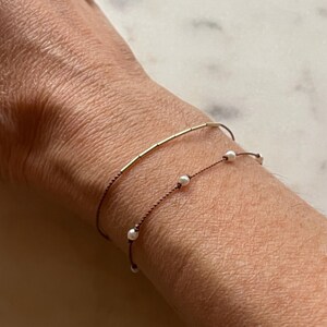May include: Two delicate bracelets on a wrist. The top bracelet is a thin gold bar. The bottom bracelet is a string with small white pearls. Both bracelets are on a brown string.