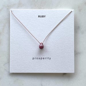 May include: A delicate necklace with a small, teardrop-shaped ruby gemstone pendant on a thin brown cord. The card reads "RUBY" and "prosperity".
