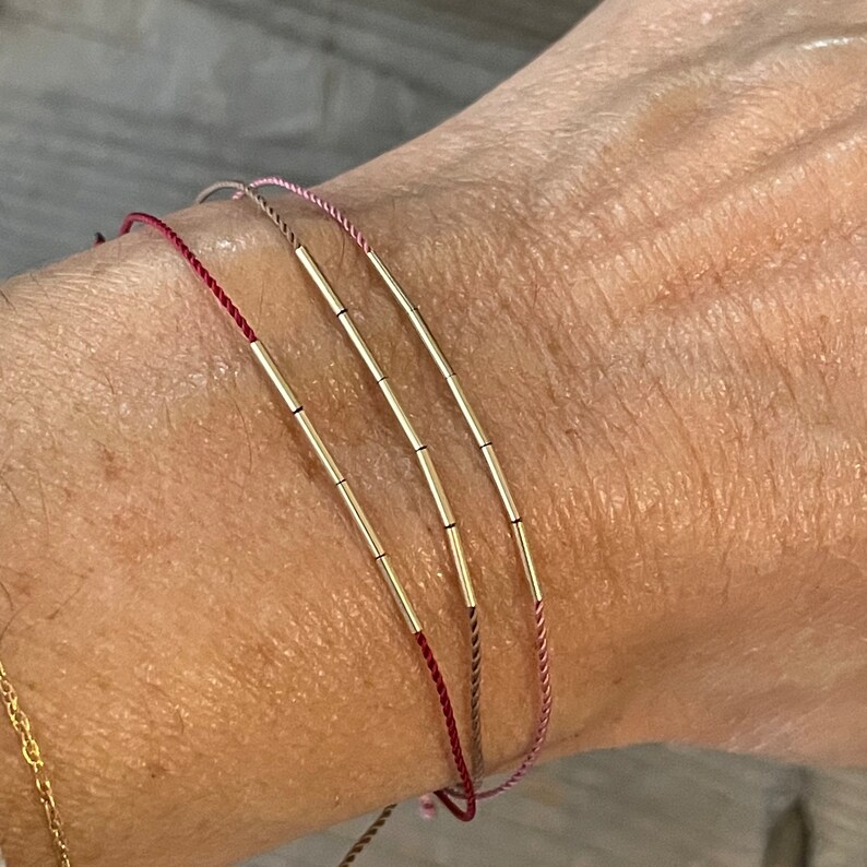 May include: Three delicate bracelets with thin, colorful cords and gold-colored cylindrical beads. The bracelets are in shades of red, pink, and tan. The bracelets are worn on a wrist.