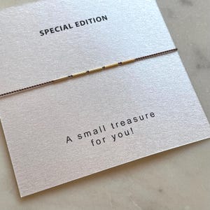 May include: A delicate gold and black beaded bracelet on a card that reads "SPECIAL EDITION" and "A small treasure for you!" The bracelet is strung on a thin, dark brown cord. The card is a light beige color.