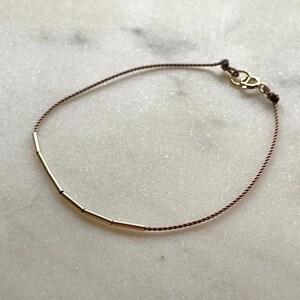 May include: A delicate bracelet featuring a thin, brown cord with a gold-colored clasp. The bracelet has several small, gold-colored cylindrical beads. The jewelry is displayed on a light-colored, marble-like surface.