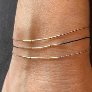 May include: Three delicate bracelets are shown, each with a thin cord and small, gold-colored cylindrical beads. The bracelets are stacked on a wrist, with one in black, one in a light brown, and one in a silver color.