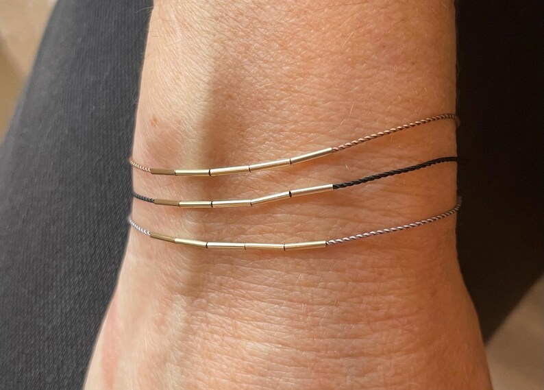 May include: Three delicate bracelets are shown, each with a thin cord and small, gold-colored cylindrical beads. The bracelets are stacked on a wrist, with one in black, one in a light brown, and one in a silver color.