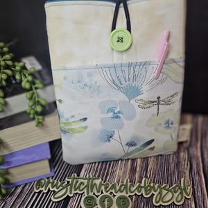 May include: A fabric book sleeve with a floral and dragonfly design in shades of blue and green. The sleeve has a pocket and is secured with a black elastic loop and a light green button. A pink pen is in the pocket.