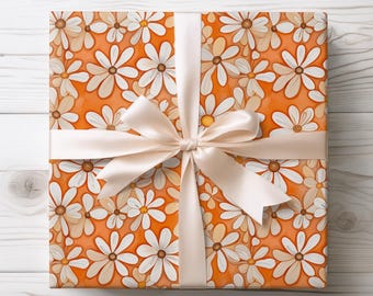 May include: A present wrapped in orange paper patterned with white daisies, tan and yellow centres. A cream satin ribbon is tied in a bow. The gift is on a wooden surface.