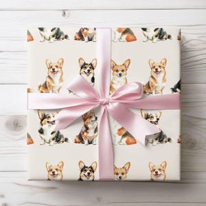 May include: Gift wrap featuring a repeating pattern of watercolor corgi dogs in various poses and colors. The gift is wrapped with a light pink satin ribbon tied in a bow. The background is a light cream color.