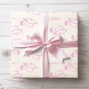 Op de afbeelding: Een witte cadeaudoos met een roze lint en een patroon van afstudeerhoeden. De doos is ingepakt in wit papier met roze afstudeerhoeden en de woorden "Gefeliciteerd" erop gedrukt.
