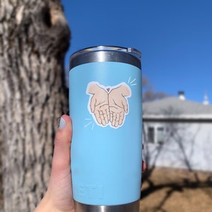 May include: A light blue stainless steel tumbler with a sticker of two hands clasped together with a small heart in the center. The sticker is white with black outlines.