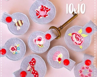 May include: Ten gray felt keychains with different embroidered designs, including flowers, a bee, a shoe, and a cupcake. The keychains have red buttons and are arranged on a pink background. The text "Schlüsselanhänger 10x10" is visible at the top of the image.