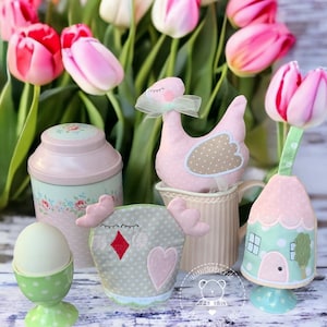 Könnte beinhalten: Ein Set aus vier pastellfarbenen Ostereierhaltern, jeder mit einem anderen Design. Die Halter sind in Form eines Küken, eines Hauses, einer Ente und eines traditionellen Eierbechers. Die Halter sind aus Stoff und haben gestickte Details. Die Halter werden auf einer weißen Holzoberfläche mit rosa Tulpen im Hintergrund präsentiert.