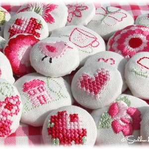 Puede incluir: Una colección de 15 botones de tela blanca con varios diseños bordados en rosa y verde. Los diseños incluyen un pájaro, una casa, un corazón, una flor y una taza.