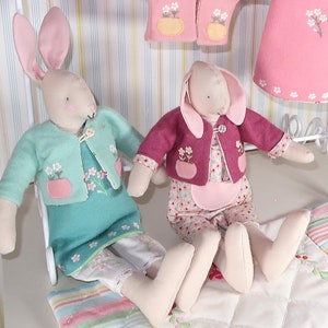 Puede incluir: Dos muñecas de peluche de conejo que llevan cárdigans rosas y verdes con bordados florales. Las muñecas están sentadas sobre una tela de rayas de colores.