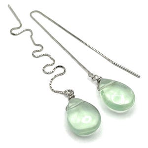 May include: A pair of silver threader earrings with teardrop-shaped, pale green gemstones. The earrings feature a delicate chain that threads through the ear, with a polished finish. The gemstones are approximately 0.5 inches in length.