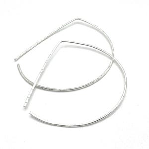 May include: A pair of silver-toned, open hoop earrings. The earrings have a hammered texture and a unique geometric design, with a straight bar that connects to a curved hoop. The earrings are displayed against a white background.
