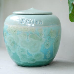 Op de afbeelding: Een turquoise en blauwe keramische urn met een deksel. Het woord "Shiloh" is in reliëf op de zijkant aangebracht. De urn heeft een kenmerkend kristallijn patroon, wat een visueel aantrekkelijk ontwerp creëert.