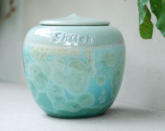 Op de afbeelding: Een turquoise en blauwe keramische urn met een deksel. Het woord "Shiloh" is in reliëf op de zijkant aangebracht. De urn heeft een kenmerkend kristallijn patroon, wat een visueel aantrekkelijk ontwerp creëert.