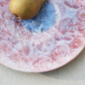 Op de afbeelding: Een rond keramisch bord met een roze en witte glazuur. De glazuur heeft een uniek, getextureerd patroon dat lijkt op kleine, witte bloemen. Het bord heeft een blauwe rand.