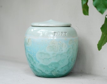 Op de afbeelding: Een keramische urn met een deksel, met een kleurengradatie van lichtblauw naar teal. Het woord "RORY" is op de urn gegraveerd. Het oppervlak heeft een textuur, kristallijn patroon. De urn is ontworpen voor decoratieve of herdenkingsdoeleinden.